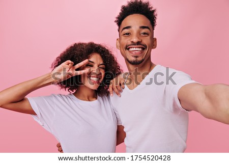 Curly woman showing peace sign and her boyfriend in white T-shirts taking selfie. Pretty brunette girl and happy man widely smile on pink background