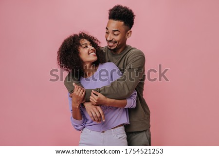 Couple in love looking at each other with love and smiling on pink background. Charming man in brown shirt hugs pretty curly woman in purple outfit