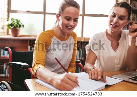 Teenage girl sitting in a wheelchair writing in a notebook with a female teacher sitting by at school. Disabled female student studying with tutor. Royalty-Free Stock Photo #1754524733