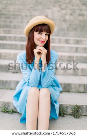 Beautiful young caucasian woman in stylish straw hat and elegant blue dress smiling, posing at camera while sitting on city stairs outdoor
