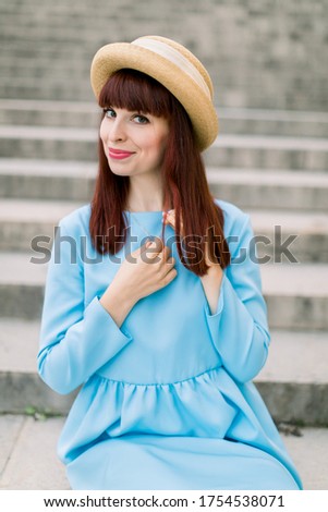 Beautiful young red haired woman wearing blue dress and hat, posing to camera while sitting on city stairs outdoors. Woman on stairs in old European city