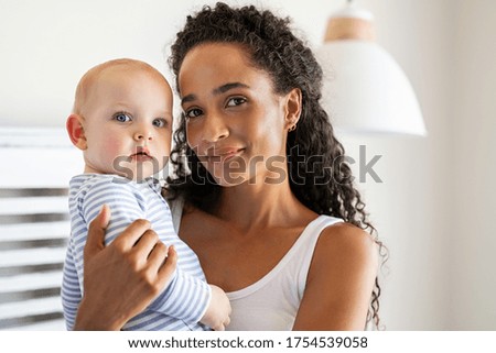 Smiling african mother holding adopted baby at home. Portrait of proud young woman rocking infant while looking at camera. Beautiful mixed race babysitter carrying adorable little child at home. Royalty-Free Stock Photo #1754539058