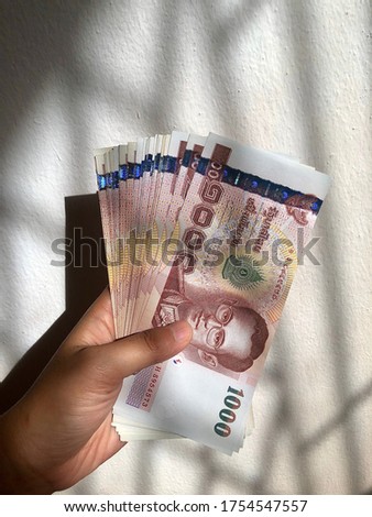Hand holding pile of Thai Baht money in bright sunlight in white stone background