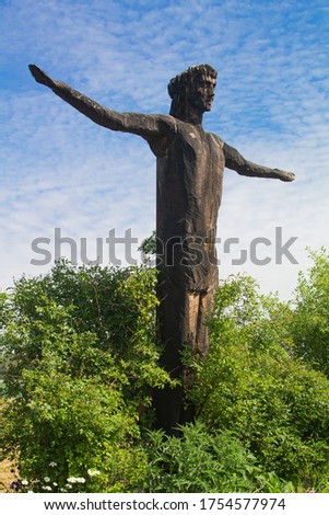 Side view picture of a wooden statue of Jesus who is standing in full pose with spread arms with a blue sky in the background