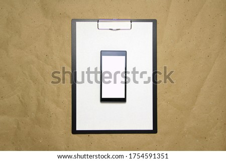 The tablet with a clip for the clip of paper with a white sheet a4 lies against the background of craft brown crumpled paper. Above is a mobile phone with a white screen. Place for text and layout for
