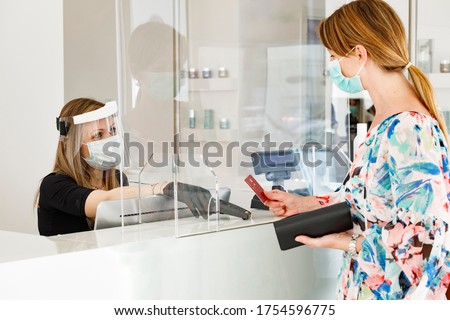 blonde girl with mask pays at the checkout with a credit card where the cashier is protected by a plexiglass plate and a visor with mask Royalty-Free Stock Photo #1754596775