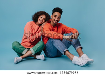 Young man and woman smiling and sitting on blue background. Portrait og guy in red sweatshirt and woman in gree jeans chilling on isolated