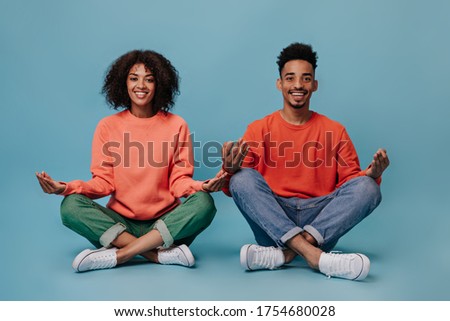 Positive guy and his girlfriend meditate on isolated background. Charming curly woman in green jeans and dark-skinned man in red sweater smile on blue backdrop