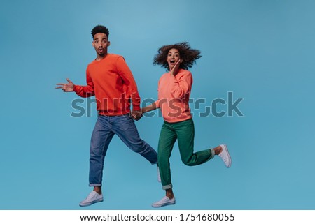 Man and woman look surprised into camera and run on blue background. Brunette girl in green pants and man in red sweater move on isolated