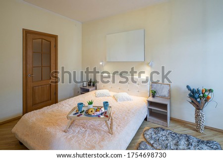 Modern interior of luxury bedroom in apartment. Light bed and bedsides with decorations. Sweets and mugs on a tray.
