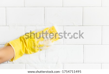 A female hand in a yellow latex glove sponges a tile. There is a lot of foam in the sink. The concept of cleaning the bathroom or kitchen, cleanliness. Copyspace. Royalty-Free Stock Photo #1754711495