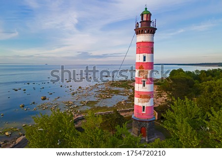The lighthouse, painted in white and red on the Bay, surrounded by greenery in the rays of the warm summer sunset in a quiet and calm weather. Lighthouse, background, landscape, nature, beauty, water.