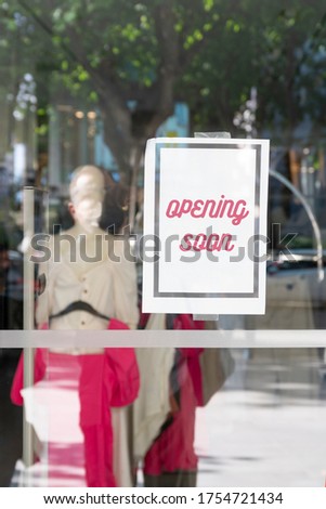 After corona virus lock down: boutique announcing OPENING SOON on a paper hanging at window. Glass reflecting environment. White mannequin inside with pink dress