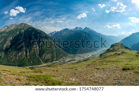 Panoramic photo of a mountain valley, a village is visible below between the mountains, clouds are floating in the sky and the mountains are covered with greenery, there is warm sunny weather.