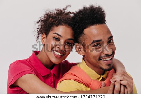 Portrait of hugging man and woman in isolated background. Cool dark-skinned girl in red shirt ans guy in yellow tee smile on white backdrop