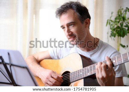 Detail of smiling man playing a song on a guitar looking at a score on a music stand at home