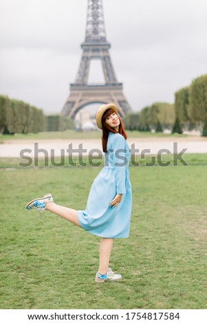 Portrait of a young excited smiling woman in straw hat and blue dress posing with one leg up,standing on green grass in front of the Eiffel tower in Paris