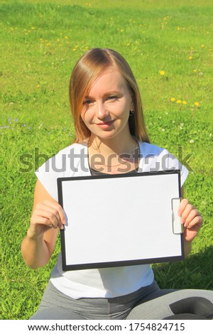 A beautiful red-haired girl in a white T-shirt sits on the green grass on the lawn and holds a white sheet of paper a4 in a black tablet with a clip. A layout for design, a place for text. High