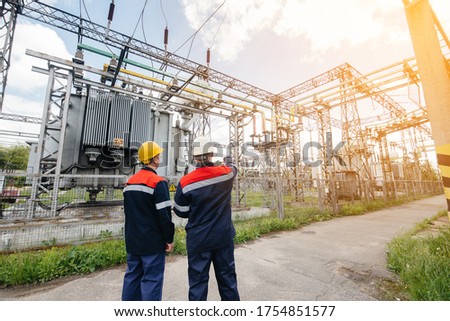 Two specialist electrical substation engineers inspect modern high-voltage equipment during sunset. Energy. Industry Royalty-Free Stock Photo #1754851577