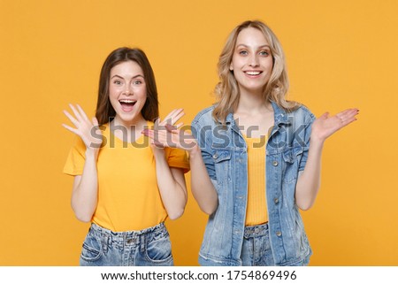 Two surprised young women girls friends in casual t-shirts denim clothes posing isolated on yellow background studio. People lifestyle concept. Mock up copy space. Keeping mouth open, spreading hands