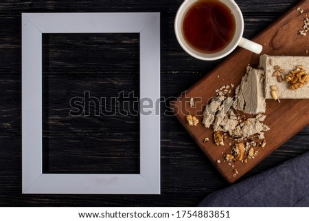 top view of an empty picture frame and halva with sunflower seeds and walnuts on a wooden board and a cup of tea on rustic background