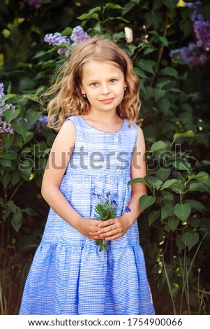 Cute little girl with blond hair and blue eyes in a summer dress. Summer portrait on a background of greenery and lilac in the garden. Image with selective focus