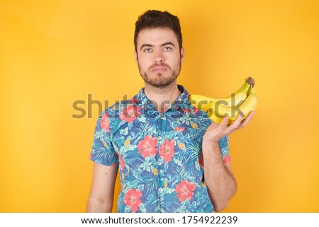 Waist up shot of joyful blue eyed boy looking to the camera, thinking about something. Holding bananas, having neutral facial expression. Standing outdoors wondering about something happening. Royalty-Free Stock Photo #1754922239