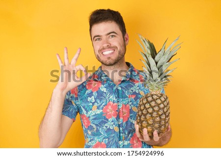 Glad attractive man shows ok sign holding an ananas on the other hand with hand as expresses approval, has cheerful expression, being optimistic. Standing against yellow wall.