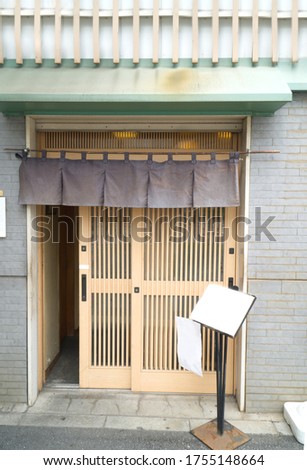 Traditional Japanese restaurants and shops style front view, curtain old fabric hangs in  front and on top of Japanese wooden slide door. The white banner paper on the stand for advertisement,