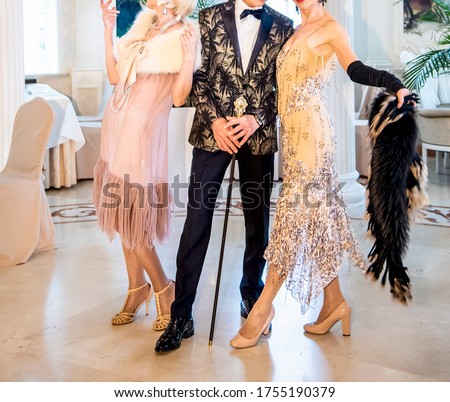 Two women in evening dresses with fur capes on their shoulders in beautiful shoes are dancing with an elegant man in a black jacket with a gold pattern and black trousers. Gatsby style party.  Royalty-Free Stock Photo #1755190379
