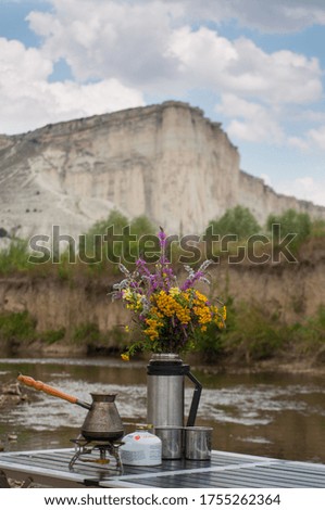 Morning at a tourist camp by the mountain river. On the folding table are camping mugs, a bouquet of wildflowers and a portable gas stove for coffee in cezve. Cosiness of camping kitchen. Copy space.