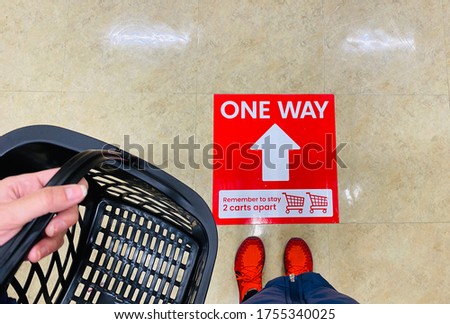 Social Distancing Sign at Store Shopper with Basket Looking Down at Ground During Pandemic COVID-19 Shopping One Way Arrow