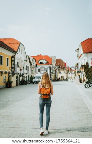Young woman traveler with long hair and backpack walks along main street of old town. Linhartov trg. Radovljica, Slovenia, Europe. Royalty-Free Stock Photo #1755364121
