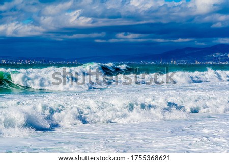 Storm waves in white foam rush in rows along the Tsemesskaya Bay. Blue sky and green sea. Dangerous and dramatic. In the background, mountains, multi-storey buildings of the city and port