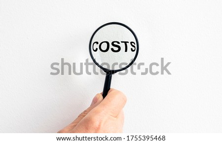 COSTS word on white wall through magnifying glass in male hands on white background