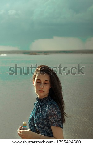 A young white woman with dark straight hair in a blue dress and with freckles stands on the background of a river on the beach. Portrait.