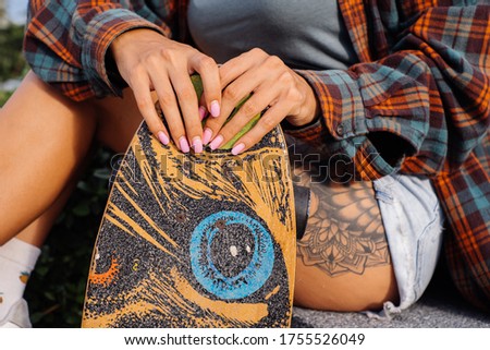 Close shot of tattooed woman in checkered shirt and jean shorts with pink pretty manicure hold colorful longboard, skateboard