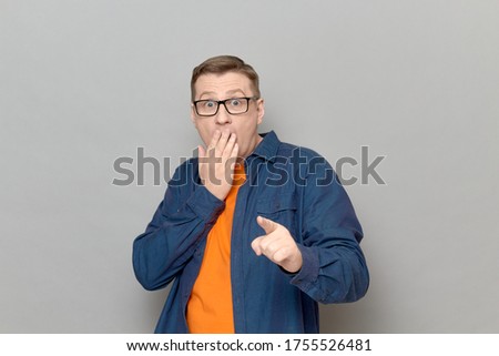 Studio shot of shocked frightened blond mature man wearing casual blue shirt, looking with eyes full of fear and pointing with finger at something awful, covering mouth with hand to prevent screaming