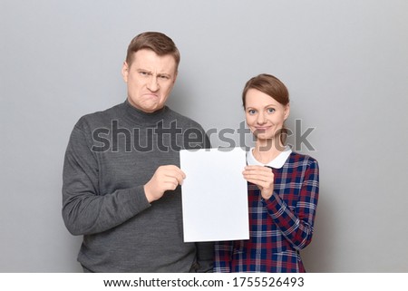 Studio portrait of adult couple holding together white blank paper sheet with place for your text, man is angry and disgruntled, woman is happy and smiling, both are standing over gray background