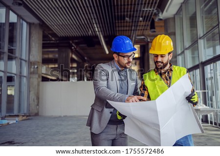 Contractor and architect looking at plan and talking about working on big project. Building in construction process interior. Royalty-Free Stock Photo #1755572486