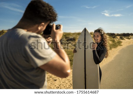 A selective focus shot of a photographer taking pictures of an attractive female holding a surfboard in the middle of the road in Spain