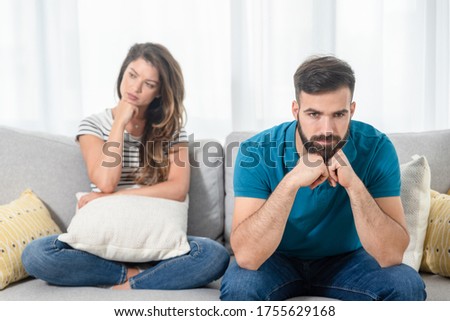 Young unhappy couple sitting on the sofa in their home in silence after arguing and fight with sad and insulting facial expressions, relationship difficulties concept selective focus