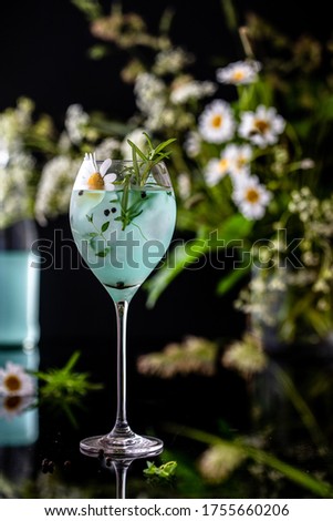 Blue gin in a tall glass covered by water droplets with summer daisy flowers as a garnish and on a background.