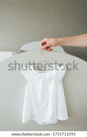 Woman’s hand holds a gold dress hanger with a simple gorgeous white dress for a child’s christening or birthday party. Pastel background, close up, front view. 
Summer wardrobe, children's clothes
