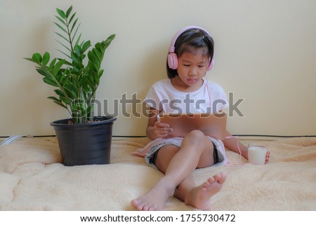 Seven years old Asian girl enjoying music with headphones and watching cartoon on tablet while holding a glass of milk at home. Children and technology, healthy food for kid concept.