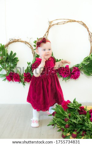 Little cute girl 1 year old in a dress of Marsala color with a wicker basket with peonies. Spring and flowers. Children's fashion