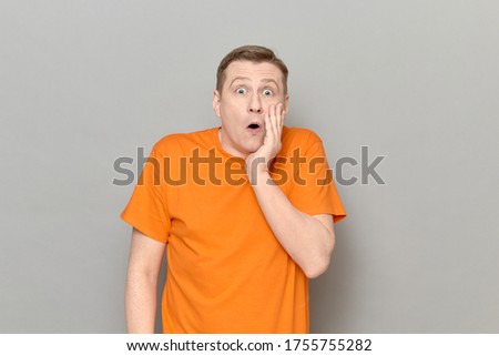 Studio portrait of shocked mature man wearing orange T-shirt, touching cheek with one hand, screaming, looking with widened eyes at something unbelievable and awful, standing over gray background
