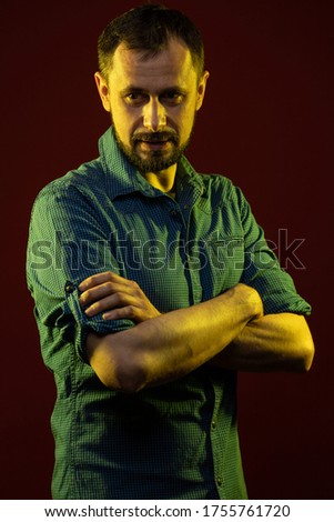 A brunette man with a beard in a blue shirt poses on a dark red background in the studio