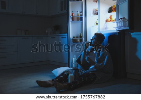 Young man eating cookies near open refrigerator in kitchen at night Royalty-Free Stock Photo #1755767882