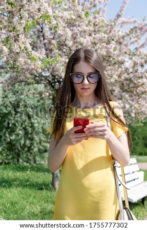 1 white young woman with long hair in a yellow dress with a phone on the background of a blooming garden